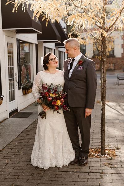 A bride and groom stand on a street, holding her bouquet of vibrant jewel tone fall colours in yellow, burgundy, deep purple and more